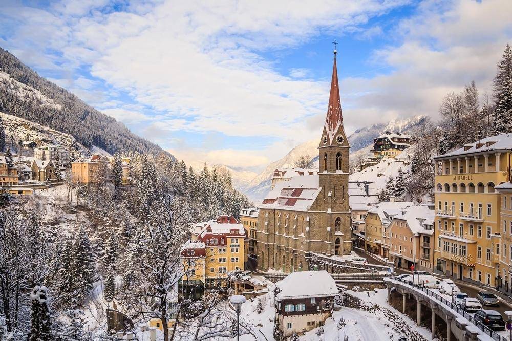 Winterlandschaft mit schneebedeckten Häusern, einer Kirche und majestätischen Bergen im Hintergrund.