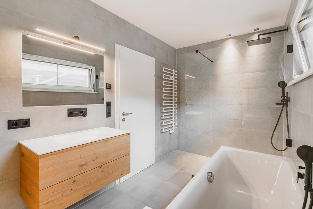 Modernes Badezimmer mit grauen Fliesen, Holzwaschtisch, Badewanne, Regendusche und Fenster.