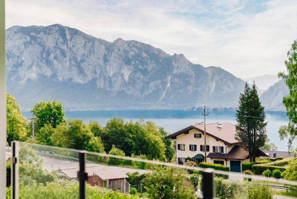 Panoramablick vom Balkon auf den See und die umliegenden Berge mit einem traditionellen Haus im Vordergrund.