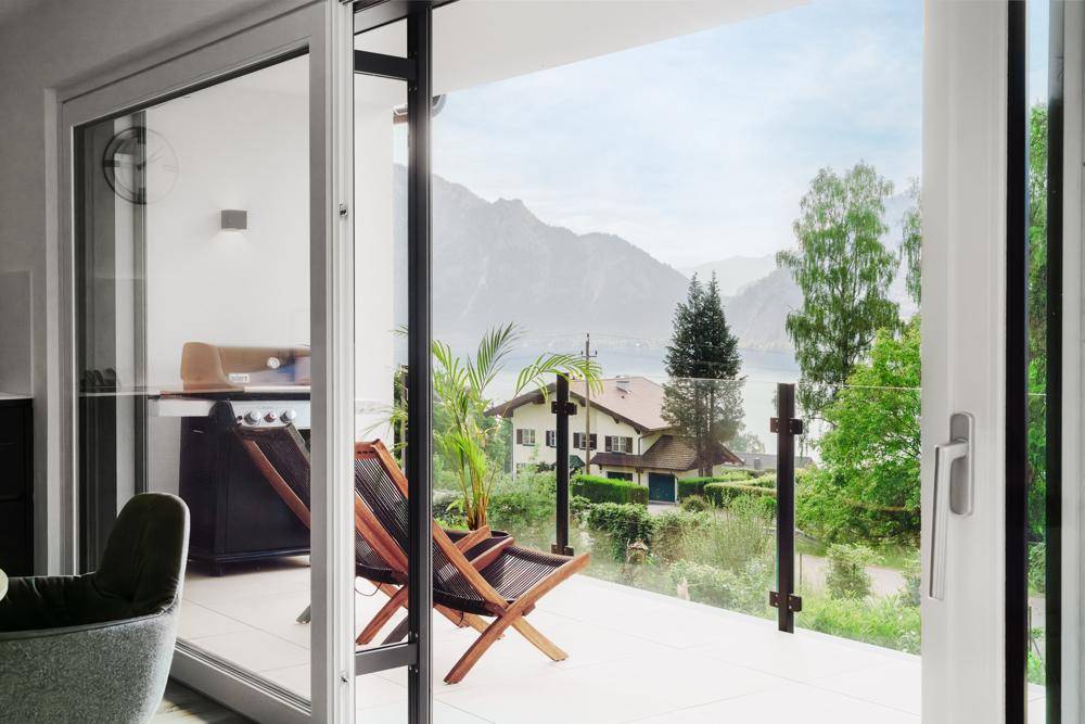 Blick vom Wohn-Essbereich durch große Schiebetüren auf die Terrasse mit Grill und die malerische Seenlandschaft.