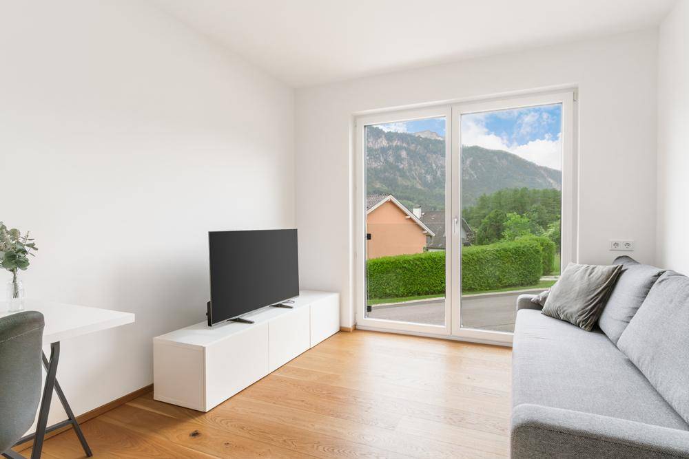 Helles Wohnzimmer mit Holzboden, gemütlichem Sofa und Blick auf die Berge und grüne Hecken.
