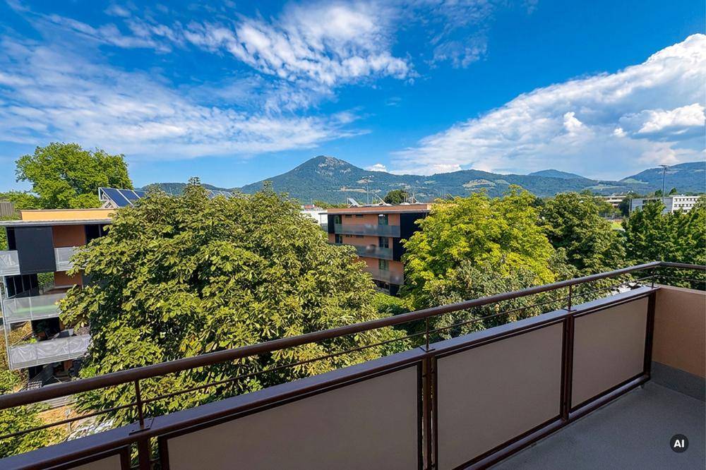 Blick vom Balkon auf grüne Bäume, umliegende Gebäude mit Solaranlagen und Berge in der Ferne.
