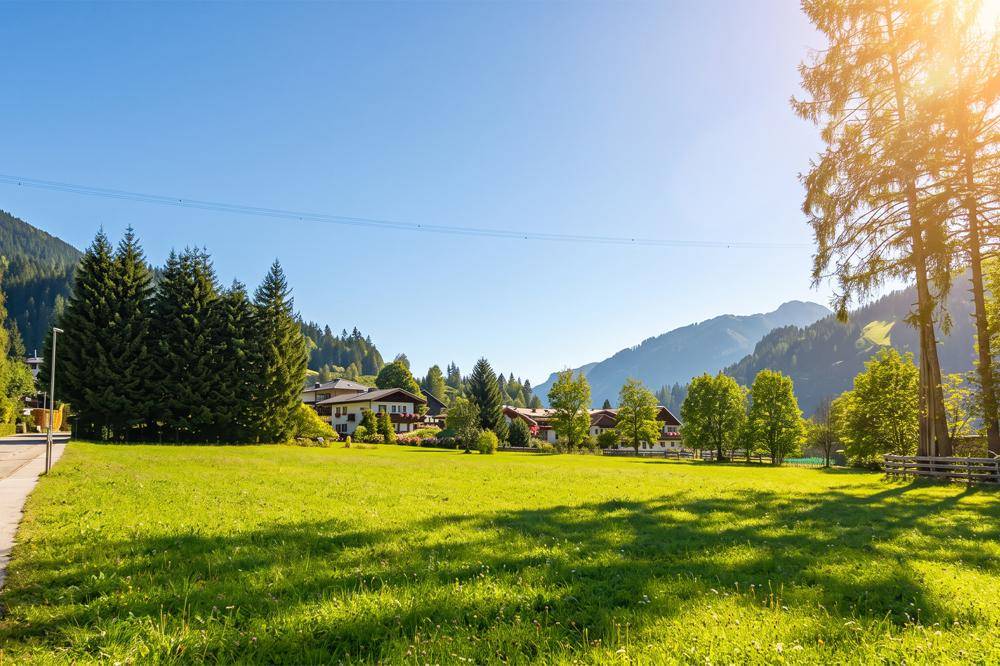 Weite grüne Wiese mit Bäumen und Häusern im Hintergrund, umrahmt von Bergen bei Sonnenschein.