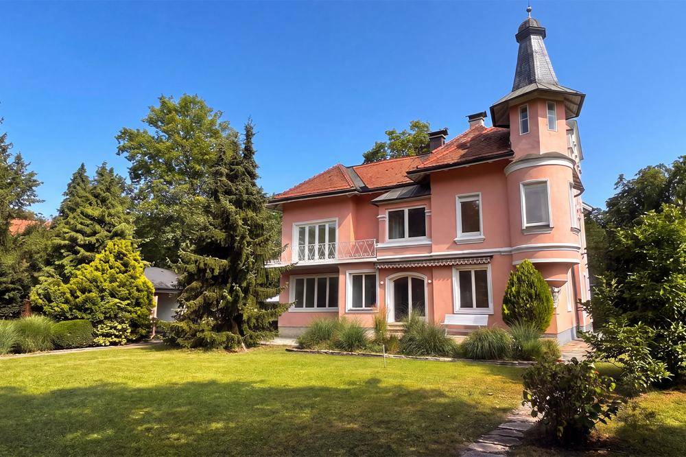Elegante rosa Villa mit markantem Turm und gepflegtem Vorgarten unter blauem Himmel.