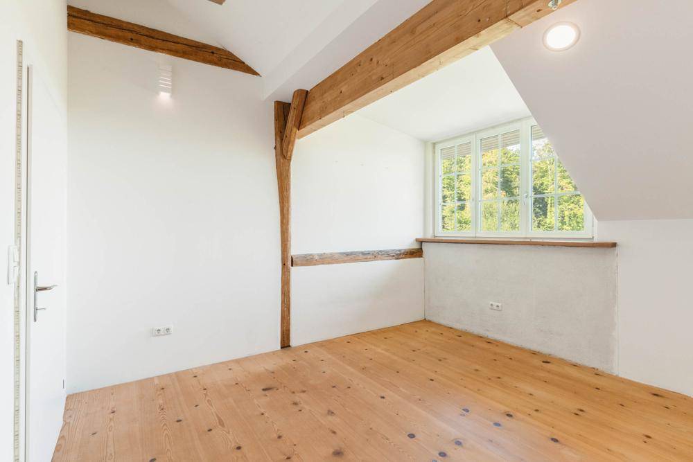 Helles Schlafzimmer mit Holzboden und sichtbaren Holzbalken, mit Blick ins Grüne.