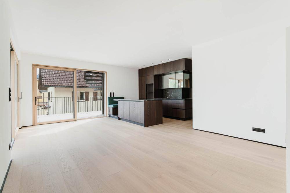 Heller offener Wohnbereich mit Holzboden, moderner dunkler Küche und Zugang zu einem Balkon.