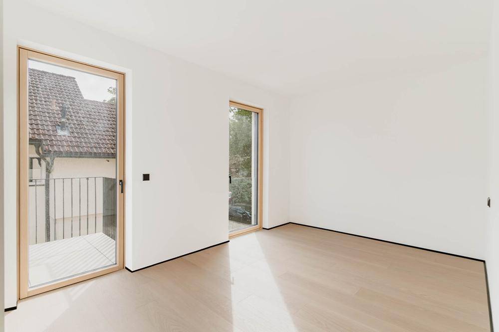 Helles, leeres Zimmer mit Holzboden und Zugang zu einem französischen Balkon und einem weiteren Fenster.