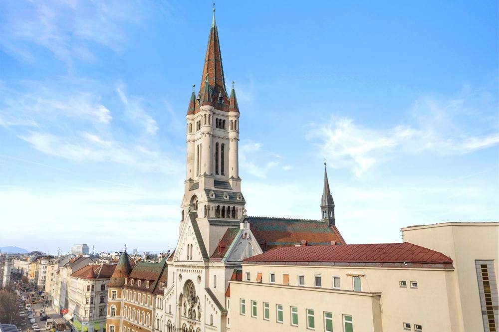 Historische Kirche mit markantem Turm, umgeben von städtischer Architektur unter blauem Himmel.