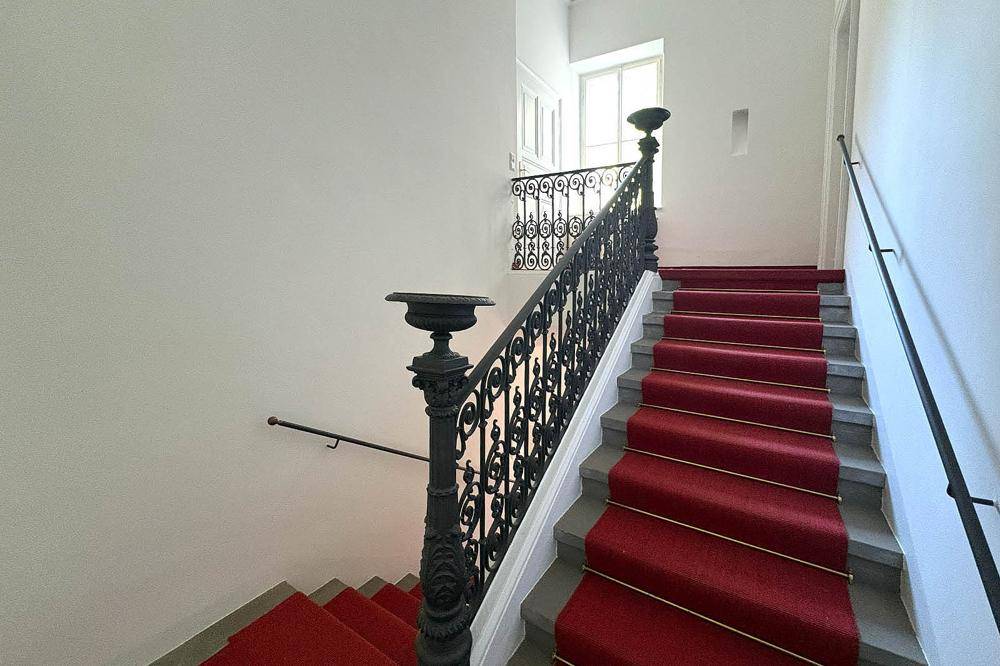 Elegantes Treppenhaus mit rotem Teppichläufer und schmiedeeisernem Geländer in einem historischen Gebäude.