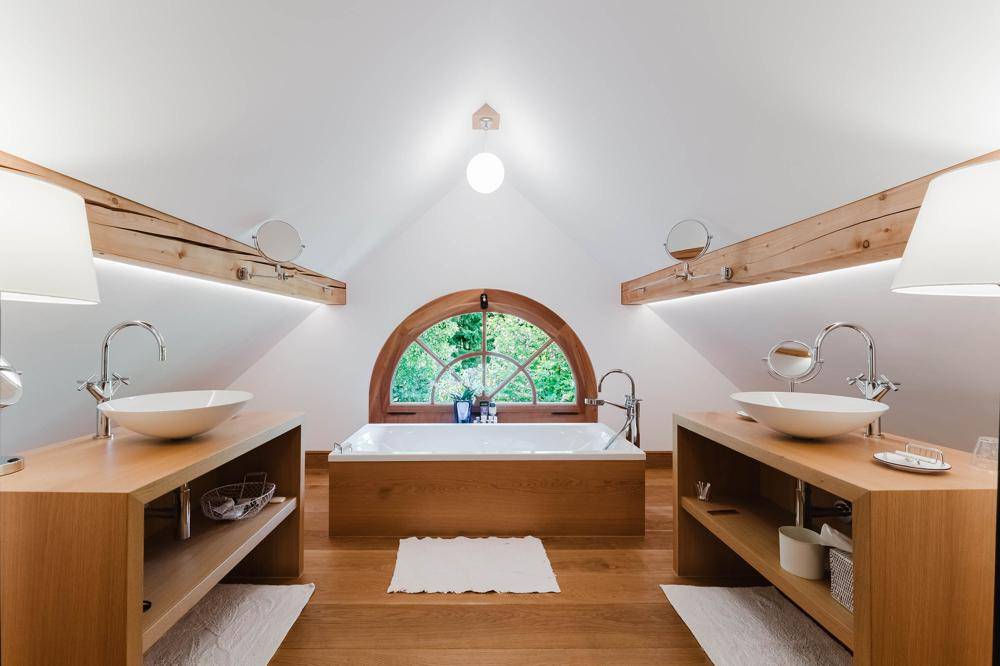 Dachgeschoss-Badezimmer mit zwei Waschbecken, Badewanne unter einem Rundfenster und Holzbalken.