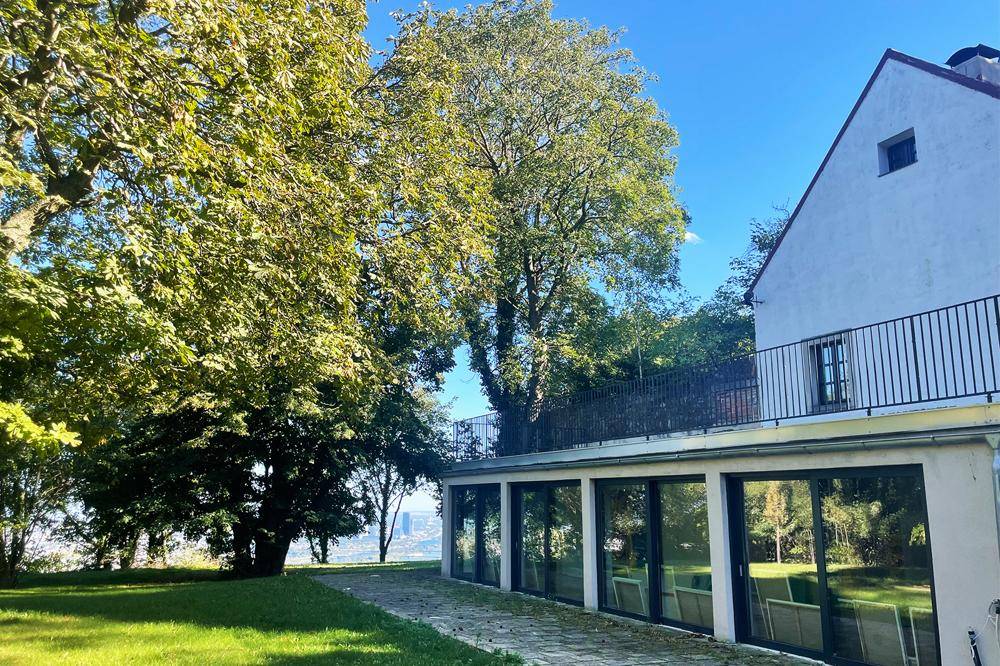 Außenansicht eines Hauses mit Glasfassade im Erdgeschoss und Balkon, umgeben von Rasen und Bäumen.
