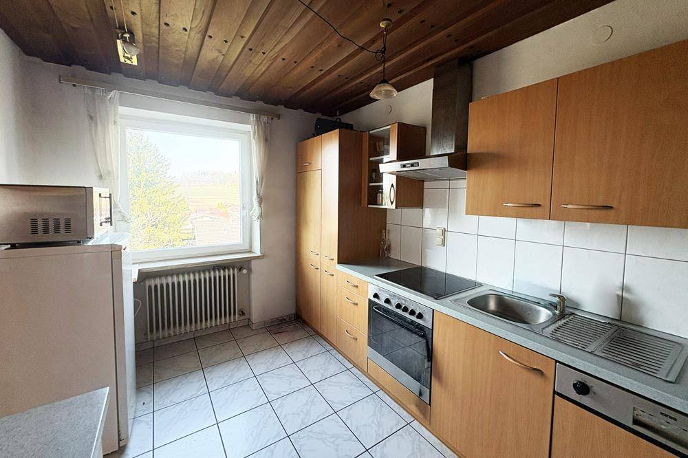 Küche mit Holzschränken, Fliesenboden und Fenster mit Blick ins Grüne, funktional eingerichtet.