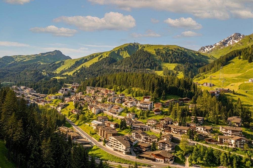 Panoramablick auf ein malerisches Dorf in den Alpen, umgeben von grünen Wäldern und majestätischen Bergen.