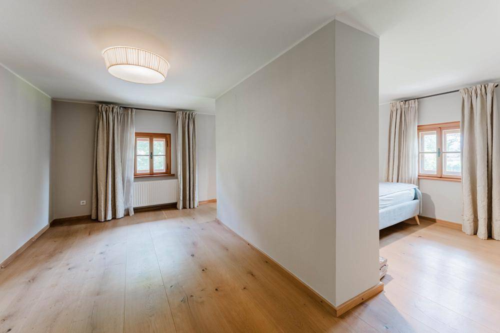 Helles Schlafzimmer mit Holzboden und zwei Fenstern, teilweise durch eine Wand getrennt.