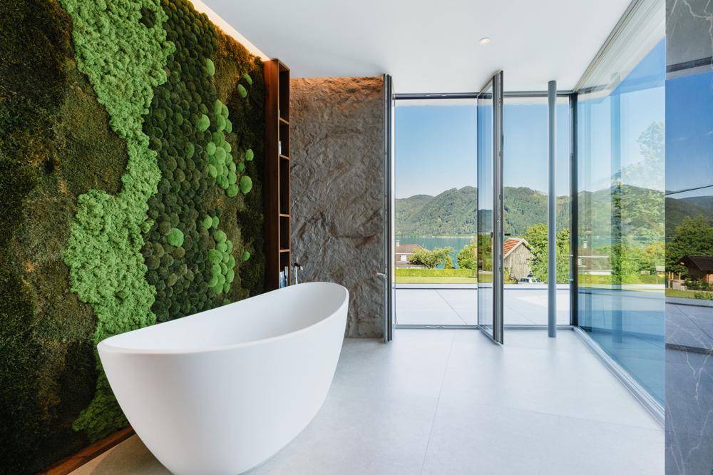 Luxuriöses Badezimmer mit freistehender Badewanne, Mooswand und Panoramafenstern mit Seeblick.
