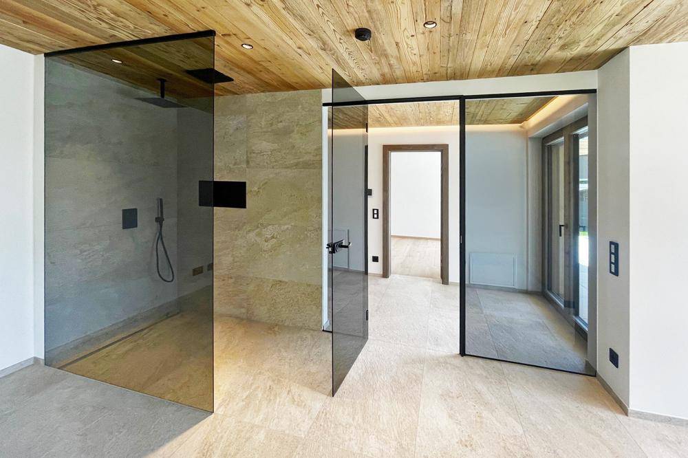 Modernes Badezimmer mit großen Glasduschen, Steinfliesen und einer Holzdecke mit sichtbaren Balken.