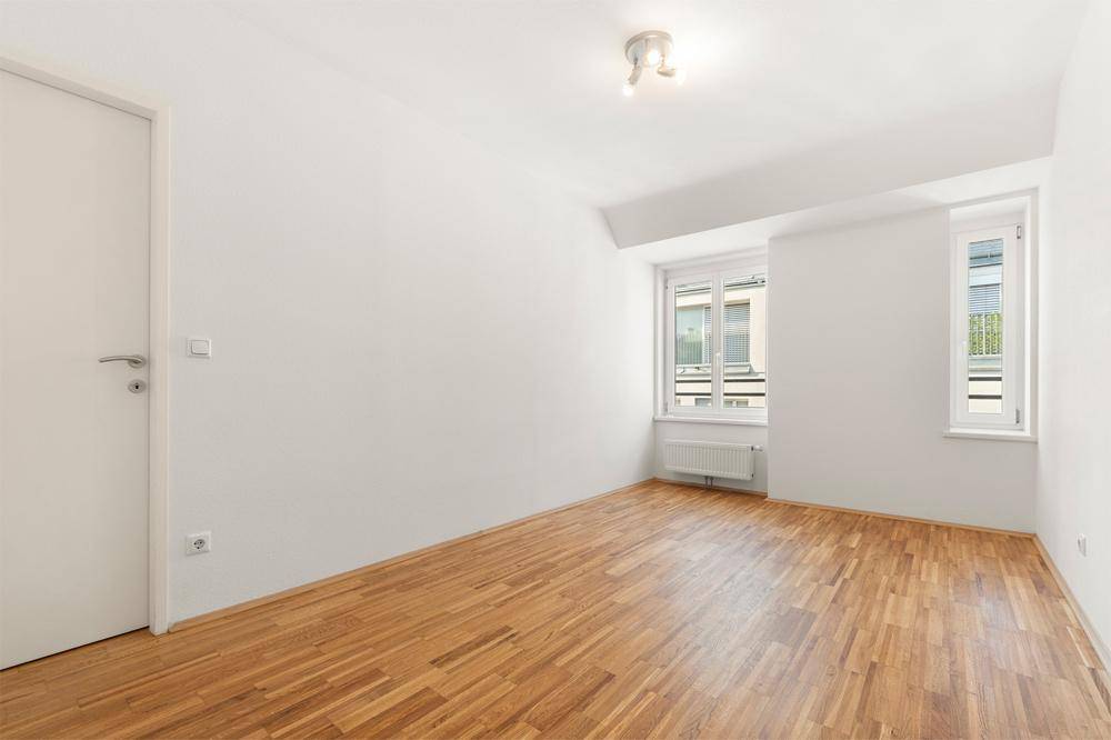 Leeres Zimmer mit hellem Holzfußboden, weißen Wänden und zwei Fenstern für Tageslicht.