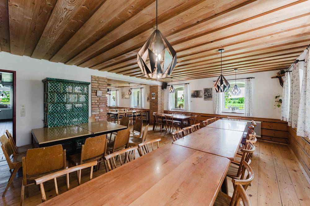 Rustikaler Speisesaal mit Holzbalkendecke, langen Holztischen und einem traditionellen Kachelofen.