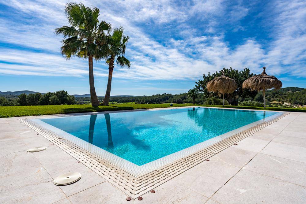 Großer, glitzernder Swimmingpool mit hellen Fliesen und Palmen unter blauem Himmel.