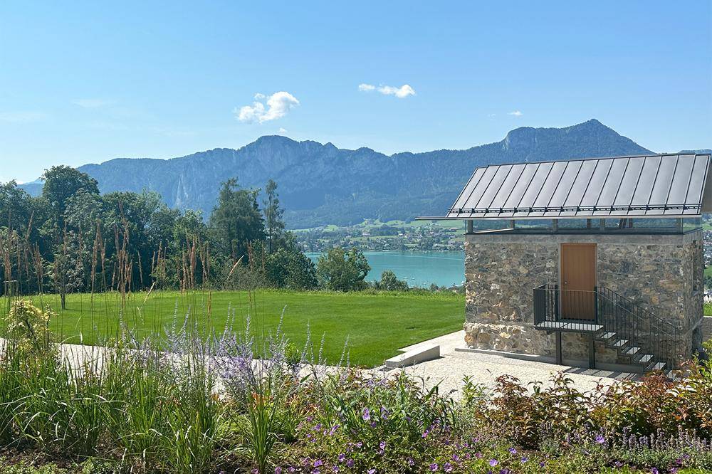 Modernes Steingebäude mit Glasdach und Treppe, umgeben von grüner Landschaft mit See- und Bergblick.