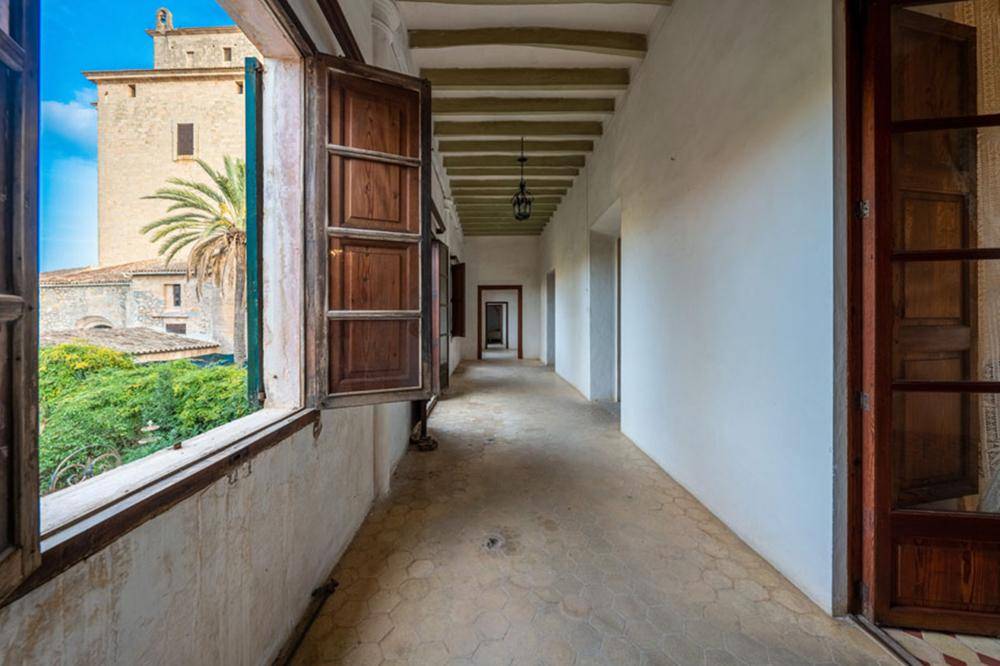 Langer Flur mit Holzbalkendecke, Fensterblick auf ein historisches Gebäude und Palmen.