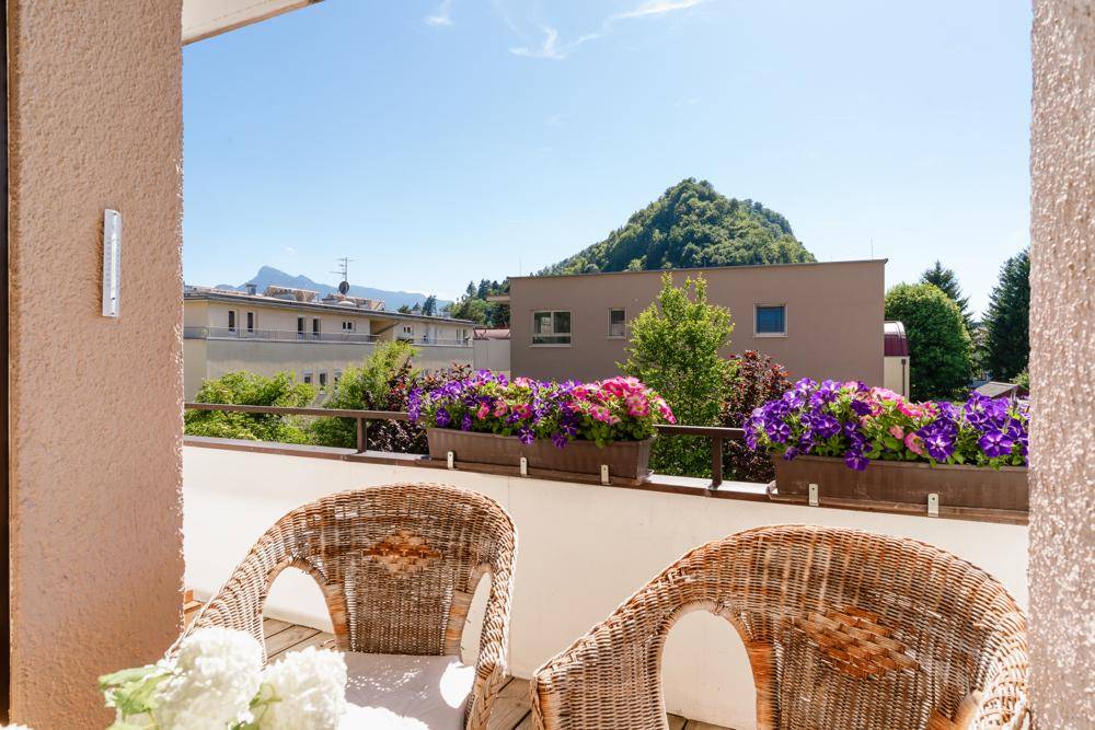 Sonniger Balkon mit Korbstühlen, bunten Blumenkästen und malerischem Bergblick.