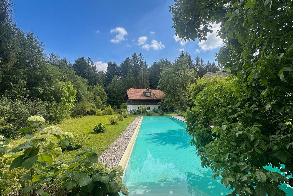 Idyllisches Anwesen mit großem Außenpool, umgeben von einem weitläufigen Garten und altem Baumbestand unter blauem Himmel.