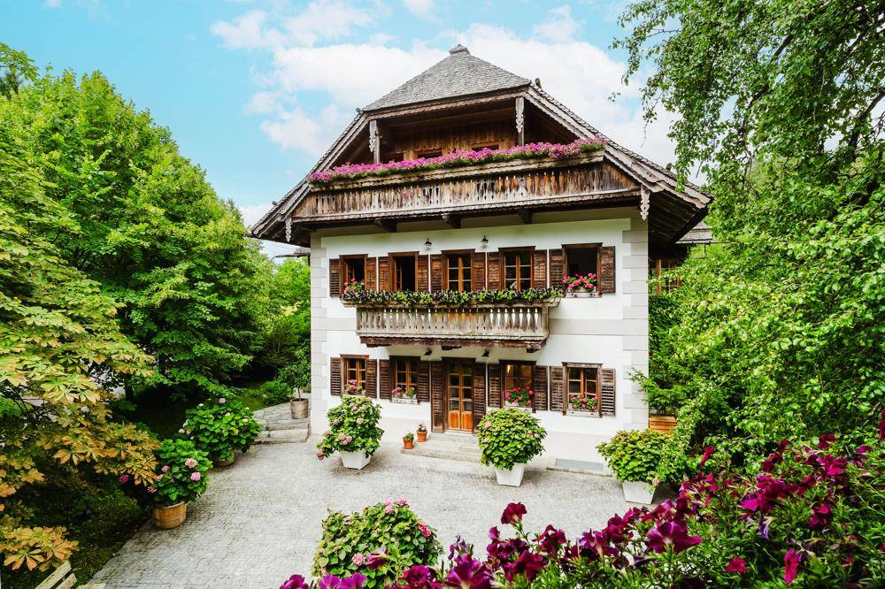 Traditionelles Landhaus mit Holzbalkonen, Fensterläden und blühenden Blumenkästen im Eingangsbereich.