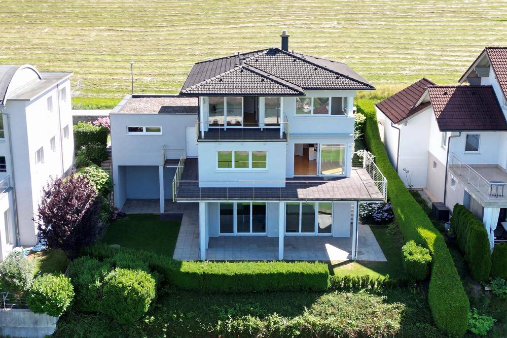 Modernes Haus mit mehreren Balkonen und Terrassen, umgeben von grünen Feldern.