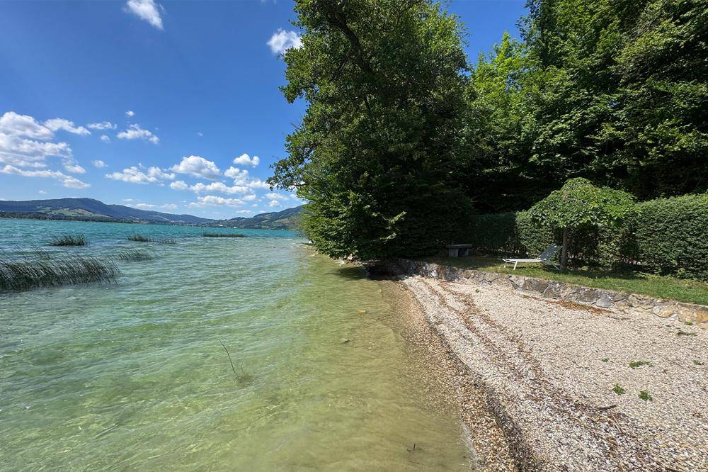 Idyllischer Seezugang mit Kiesstrand, Liegewiese und schattenspendenden Bäumen.