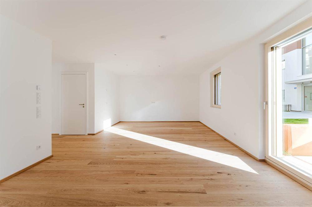 Geräumiges, leeres Wohnzimmer mit hellem Holzboden und großem Fenster, das viel Tageslicht hereinlässt.