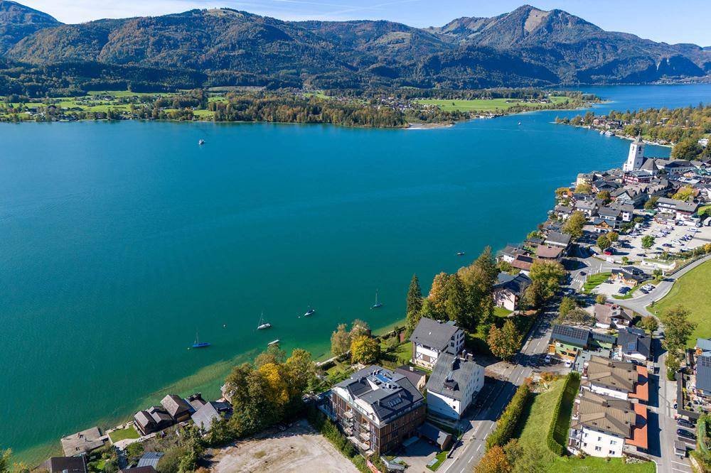 Luftaufnahme eines malerischen Sees mit umliegenden Dörfern und Bergen unter strahlend blauem Himmel.