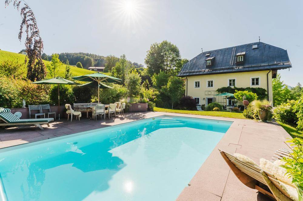 Großer Außenpool mit weitläufiger Terrasse, umgeben von grüner Natur und mit Blick auf die Berge.