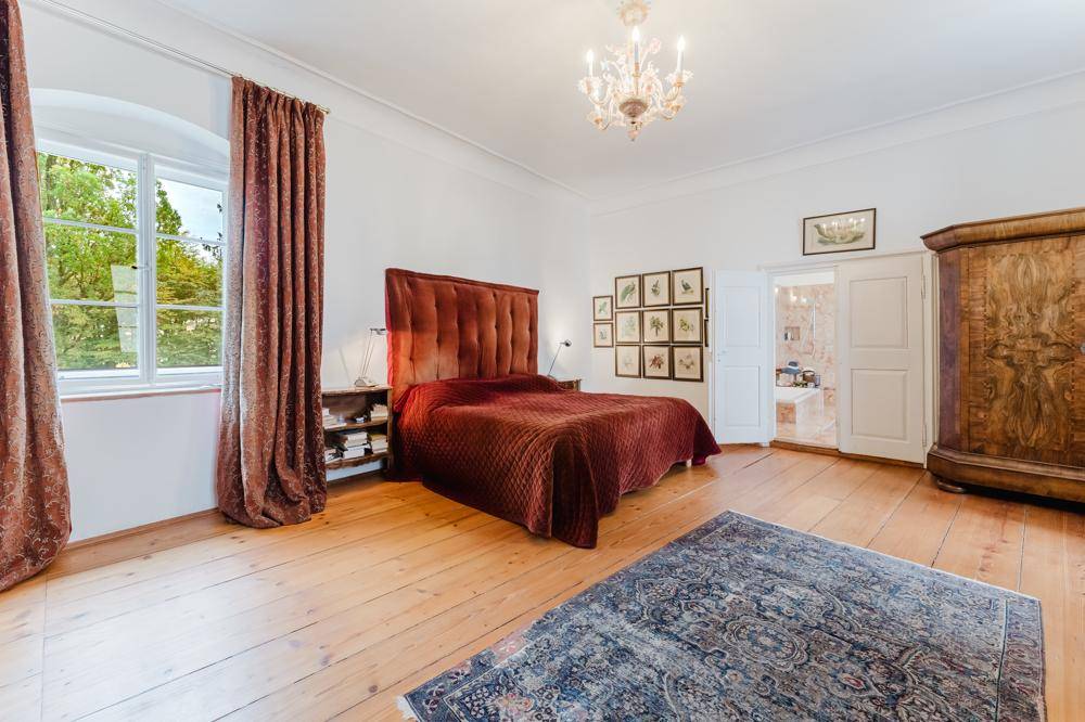 Geräumiges Schlafzimmer mit großem roten Bett, Holzdielenboden, Kronleuchter und schweren Vorhängen am Fenster.