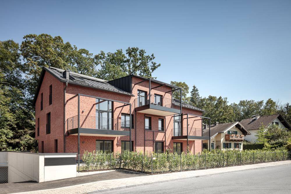 Modernes Mehrfamilienhaus mit Ziegelfassade, Balkonen und umgeben von viel Grün unter blauem Himmel.
