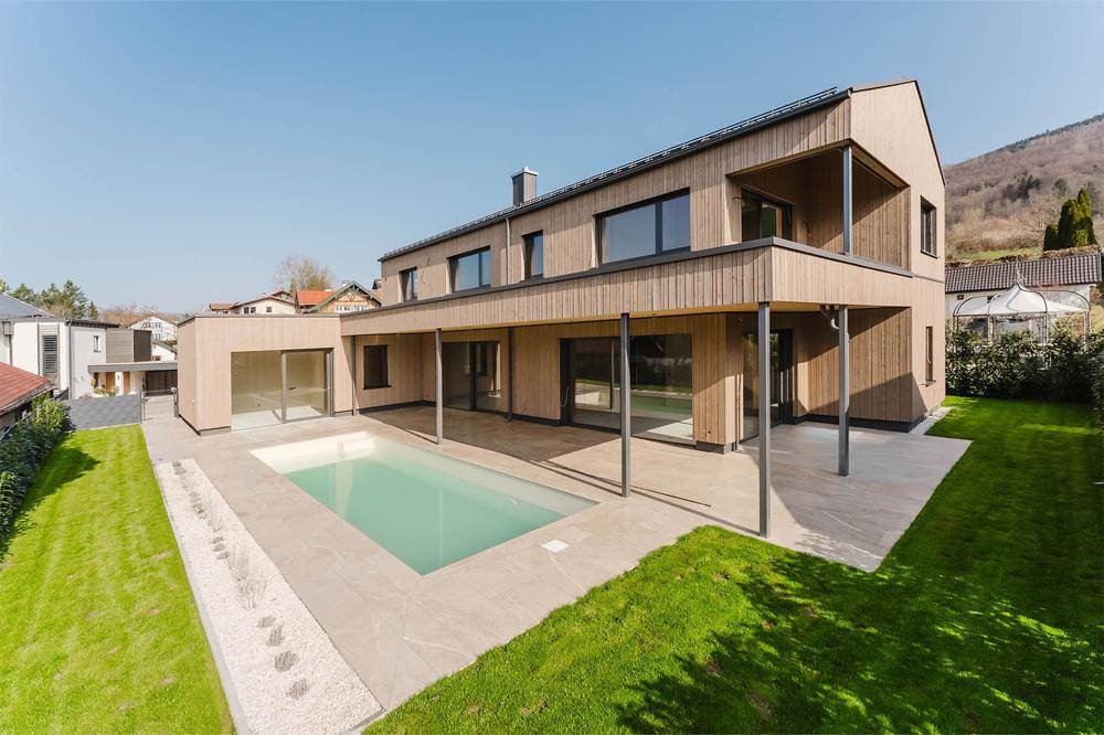 Modernes Holzhaus mit großem Pool, Rasenfläche und Bergblick unter blauem Himmel.