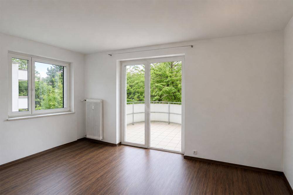 Helles Zimmer mit dunklem Laminatboden und Zugang zu einem Balkon mit grüner Aussicht.