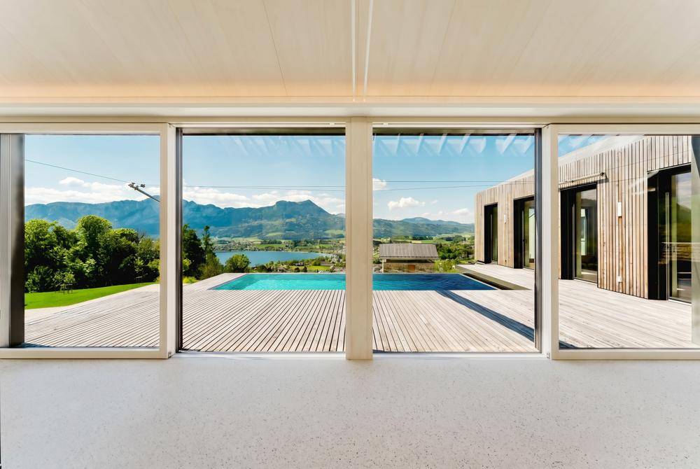 Blick aus großen Fenstern auf eine Holzterrasse mit Pool, See und majestätischen Bergen.