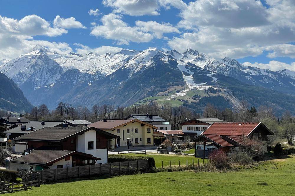 Malerischer Blick auf ein Dorf in den Alpen mit schneebedeckten Bergen und grünen Wiesen.