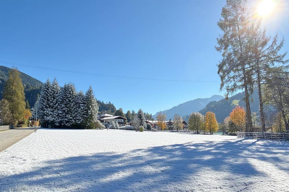 Ein weites, schneebedecktes Feld mit Häusern und bewaldeten Bergen im Hintergrund, beleuchtet von der Wintersonne.