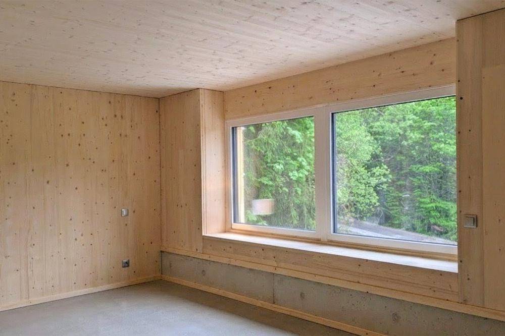 Heller, unmöblierter Raum mit Holzwänden, Betonboden und großem Fenster zur grünen Natur.