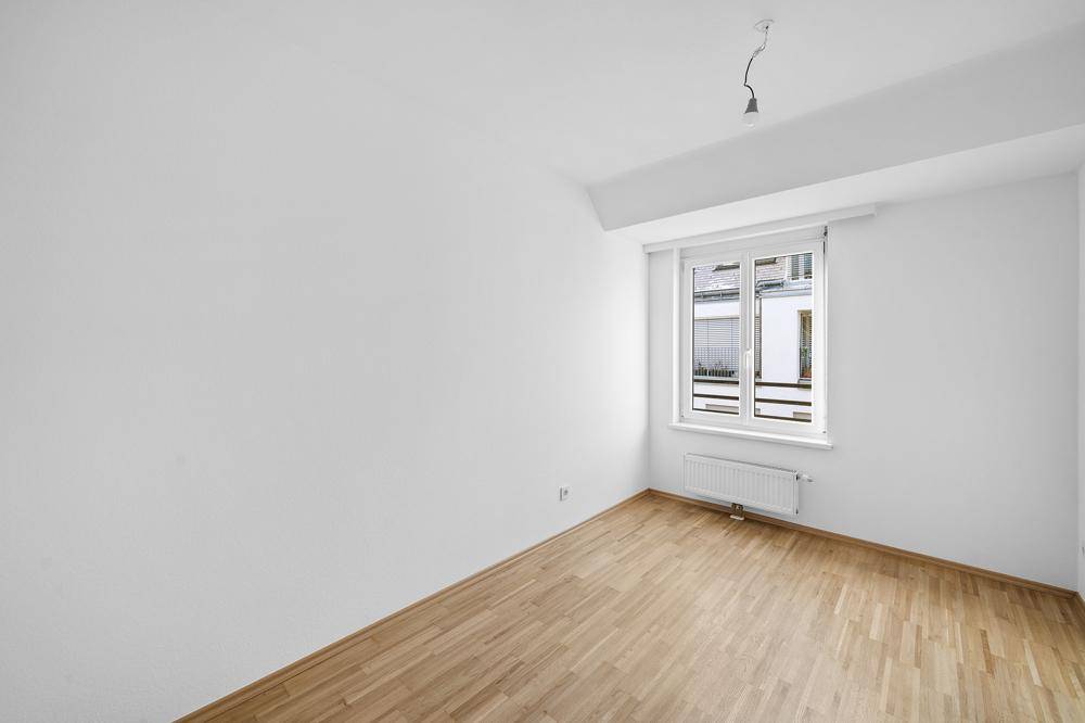 Leeres Schlafzimmer mit Holzboden, weißen Wänden und einem Fenster, das viel Licht hereinlässt.