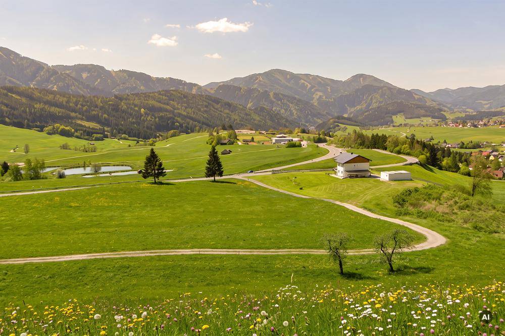 Idyllische Landschaft mit grünen Wiesen, einem Haus an einer kurvigen Straße, kleinen Teichen und Bergen im Hintergrund.