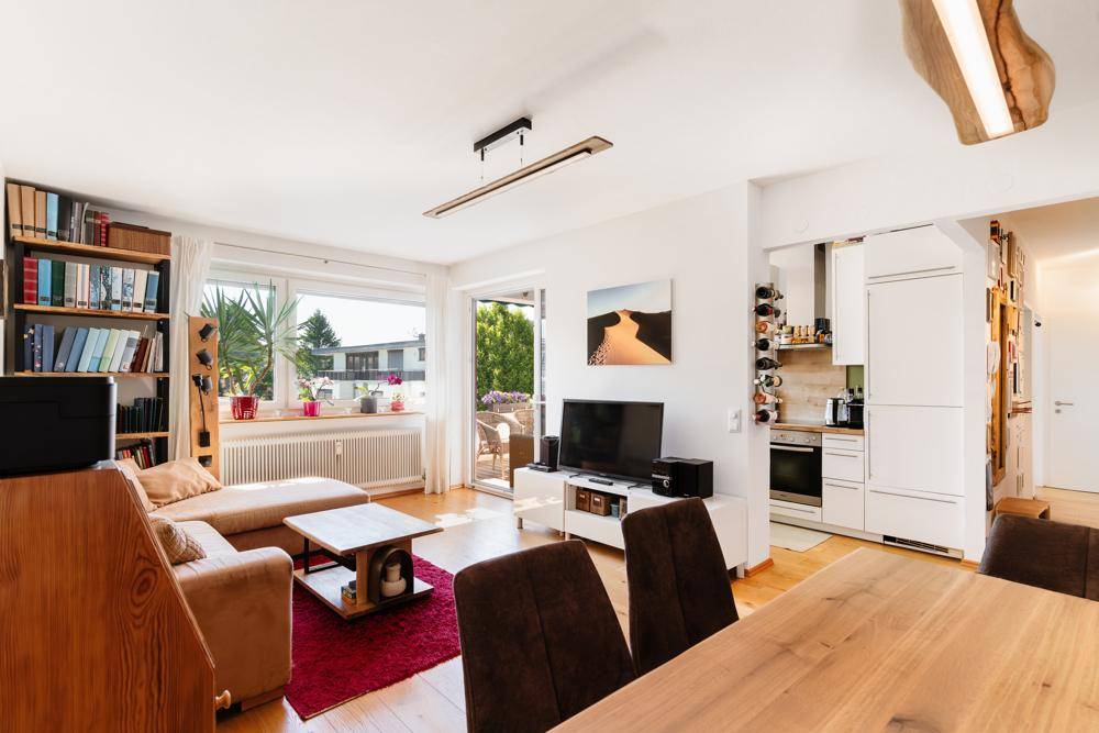 Geräumiger Wohn-Essbereich mit Holzboden, Zugang zum Balkon und offener Küche im Hintergrund.