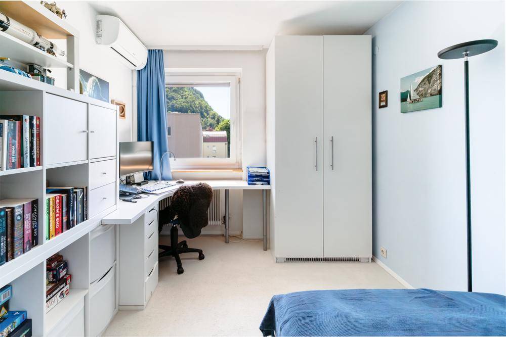 Schlafzimmer mit Arbeitsbereich, weißem Kleiderschrank, Bücherregal und Klimaanlage.