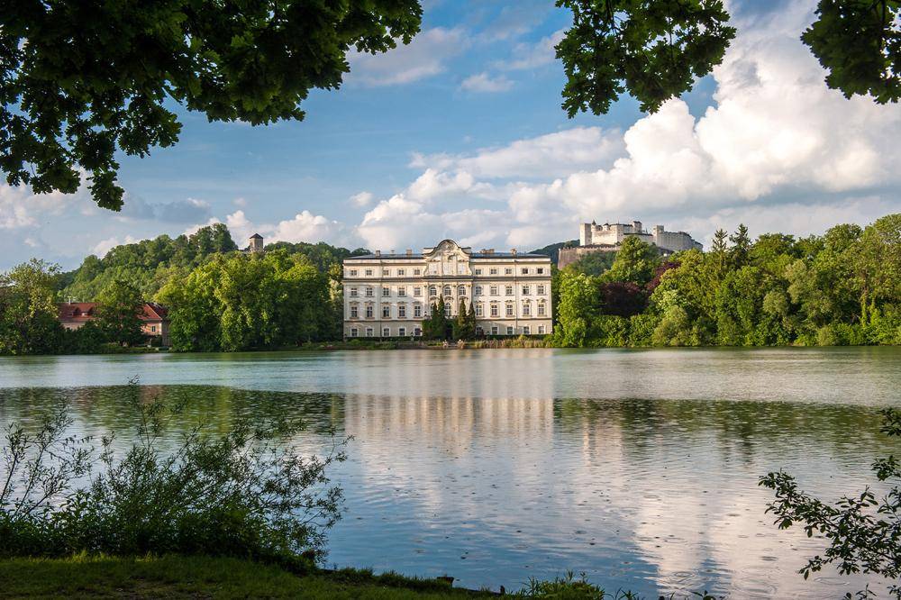 Prächtiges historisches Gebäude am Seeufer mit Spiegelung im Wasser und Burg im Hintergrund.