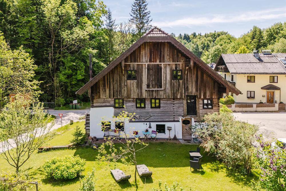 Traditionelles Holzhaus mit weißem Sockel, umgeben von einem grünen Garten und Bäumen.