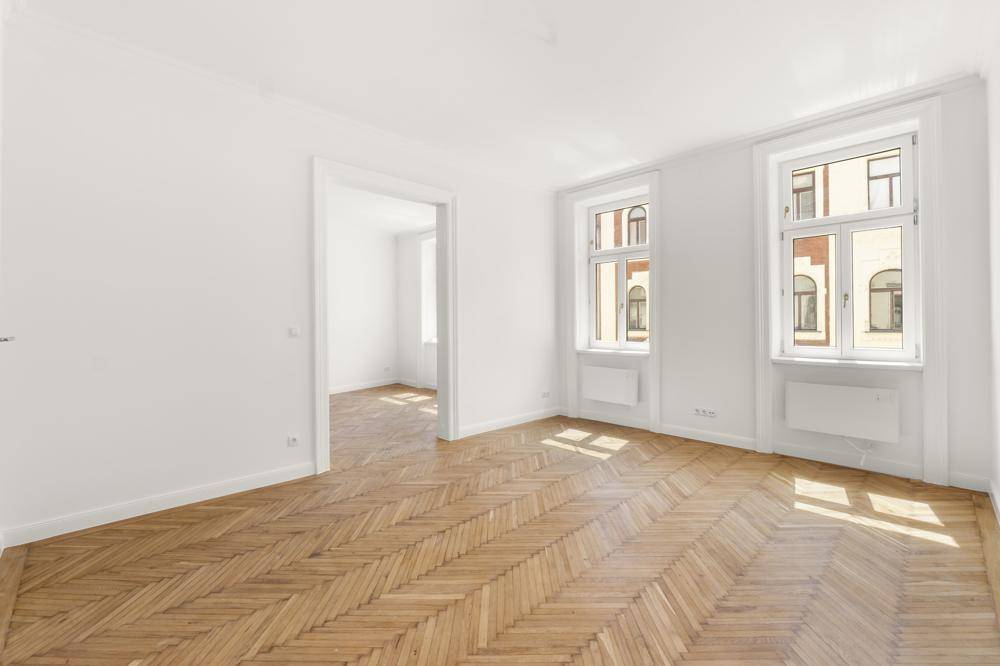 Geräumiges, leeres Zimmer mit Fischgrätparkett und hohen Fenstern, bereit zur individuellen Gestaltung.