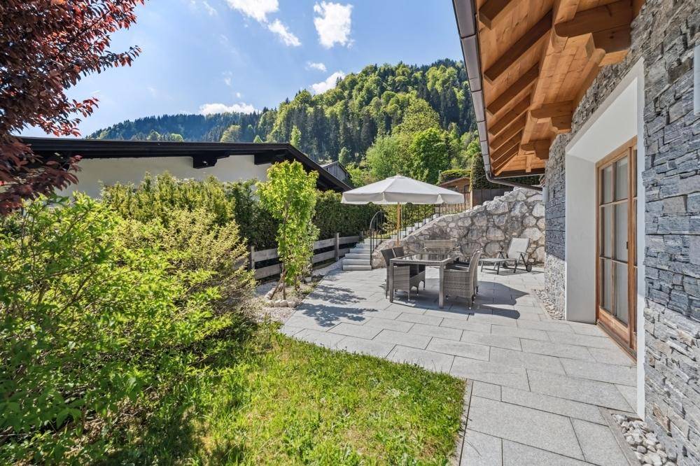 Sonniger Außenbereich mit Steinterrasse, Gartenmöbeln und Blick auf die Berge.