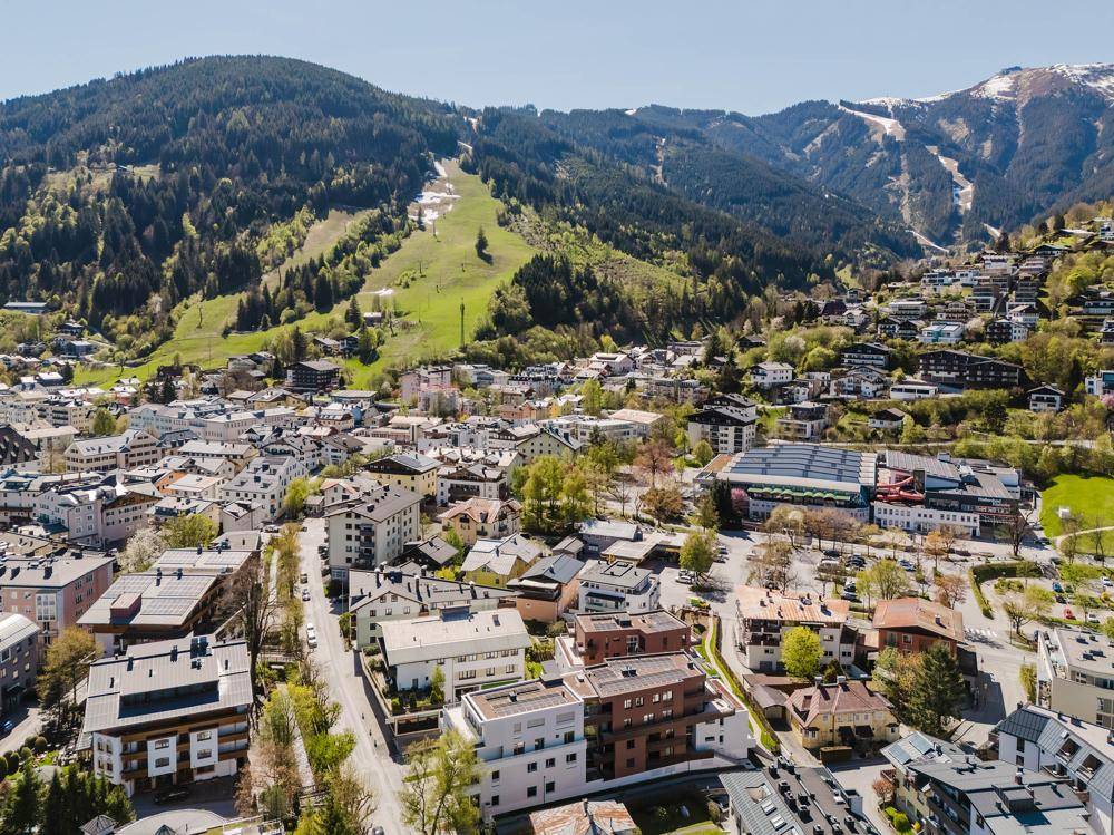 Weitläufige Luftaufnahme einer Bergstadt mit dichter Bebauung und grünen Skipisten in den Alpen.